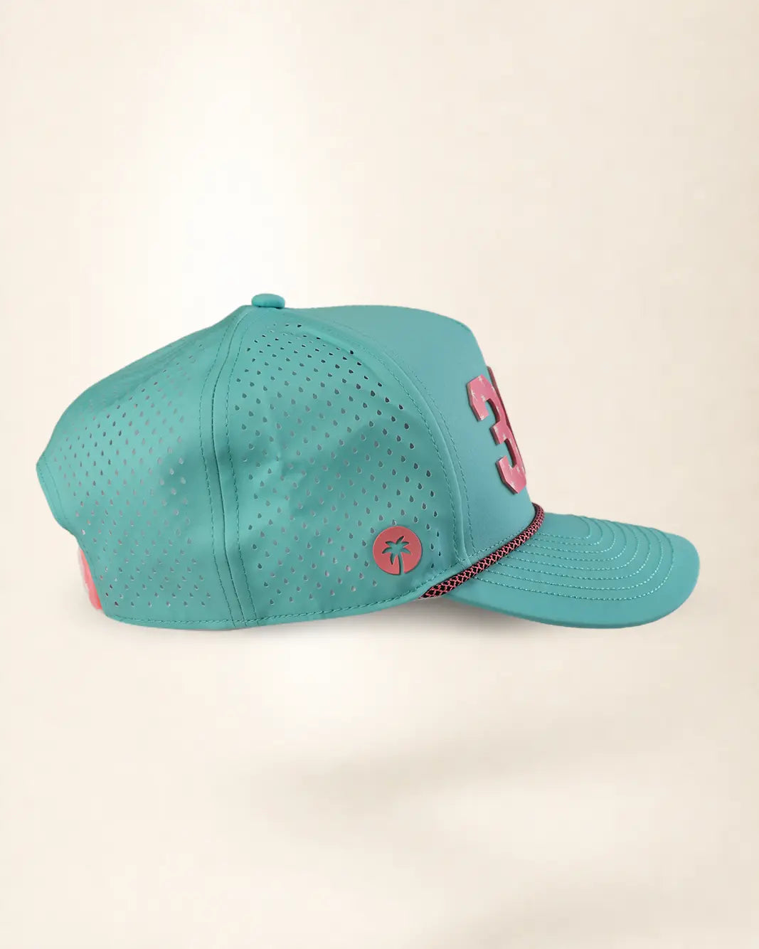 A teal perforated baseball cap with a pink embroidered number 3 on the side and a small pink circular logo patch, shown against a plain cream background.