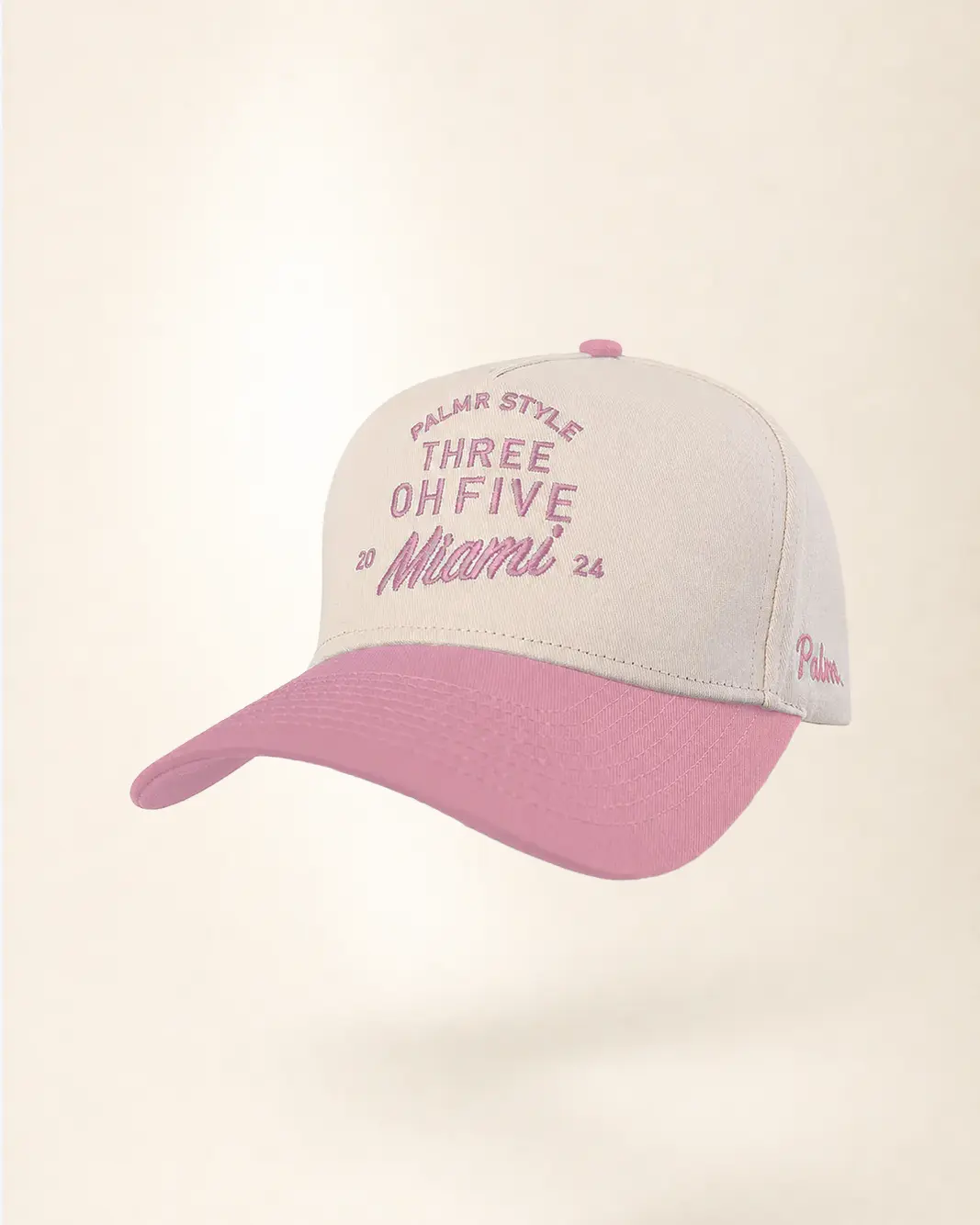 A baseball cap with a light beige crown and a dusty pink brim, featuring embroidered pink text on the front and side.