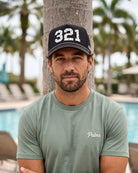 A man wearing a black baseball cap with '321' in white and a sage green t-shirt with 'Palmr' embroidered on the chest, standing in front of a palm tree and a swimming pool.