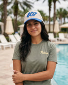 A woman standing by a swimming pool wearing a beige and blue trucker hat with '407' on it, and an olive green t-shirt with 'Palmr' embroidered in gold on the chest.