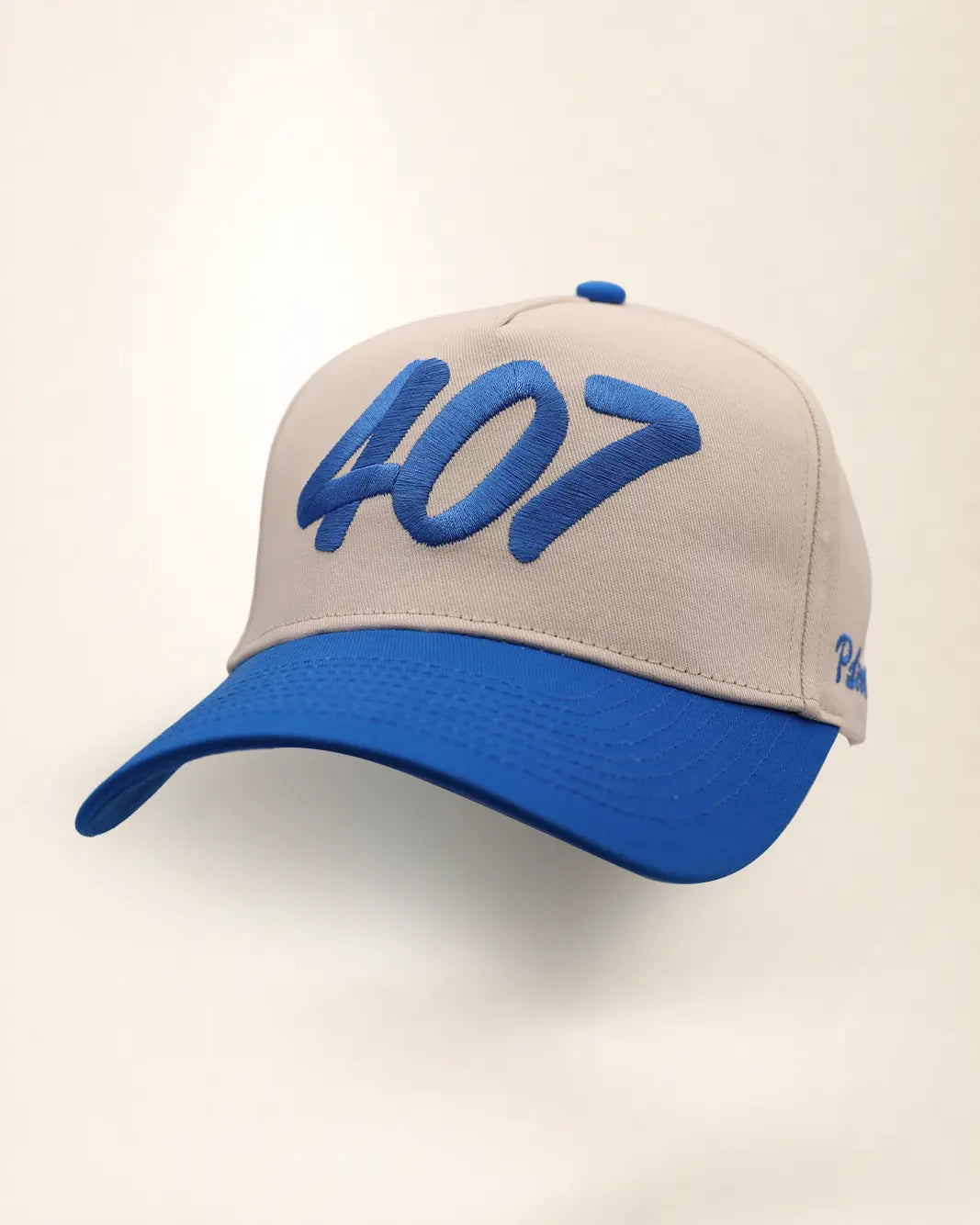 A baseball cap with a beige crown and a royal blue brim, featuring the number '407' embroidered in blue on the front. A small blue logo is visible on the side.