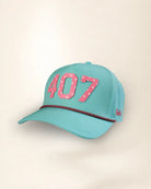 A turquoise baseball cap with '407' in pink star-patterned letters on the front, a thin red and black patterned rope detail across the brim, and a small red script logo on the side, set against a plain beige background.