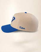 A structured baseball cap in beige and royal blue, featuring a beige crown with a blue embroidered letter 'V' on the side and the name 'Palmr' embroidered in blue script on the front. The curved brim is royal blue.