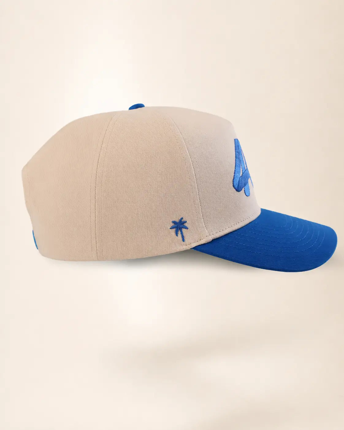 A structured baseball cap with a light beige crown and a bright blue brim, featuring a small blue embroidered palm tree on the side and a larger blue graphic on the front.