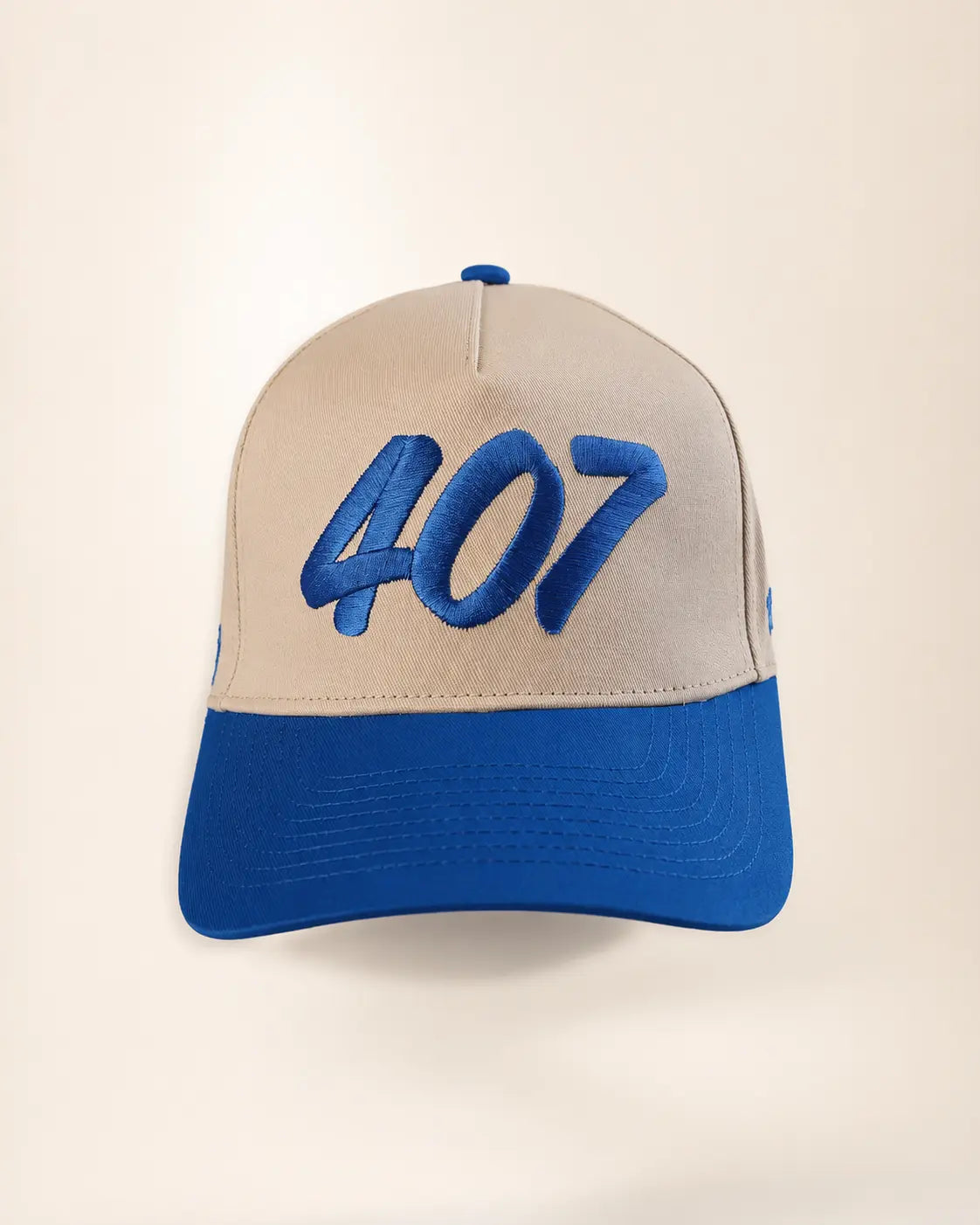 A baseball cap with a beige crown and a royal blue brim, featuring the number '407' embroidered in blue on the front. A small blue logo is visible on the side.
