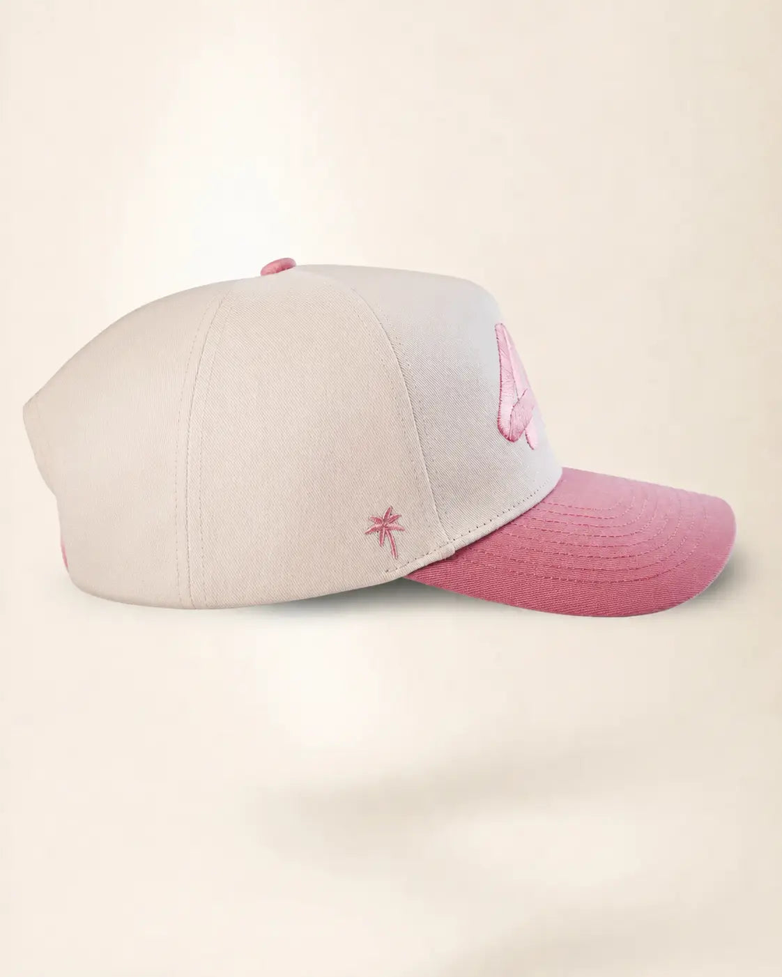A baseball cap with a light gray crown and a dusty pink brim, featuring a small pink embroidered palm tree on the side and a pink embroidered letter on the front.