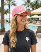 A woman in the pink 813 Signature Hat turned slightly away, with palm trees and a pool behind her.