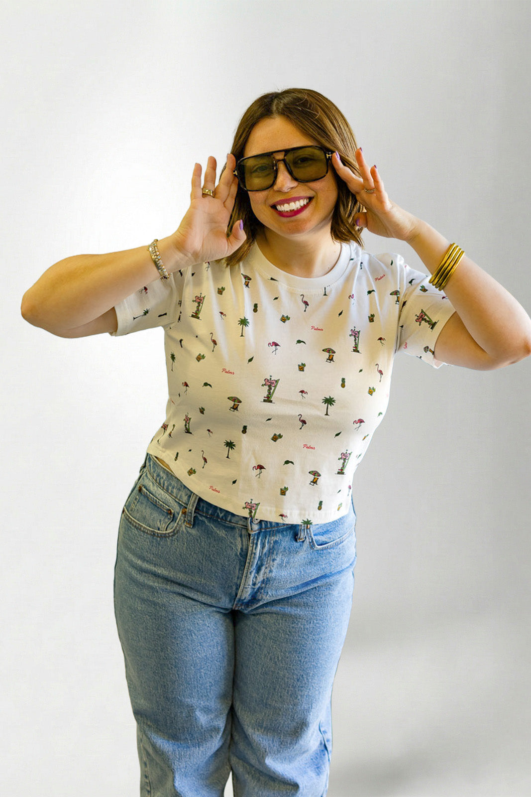 Woman wearing a patterned shirt and jeans with sunglasses on a plain background