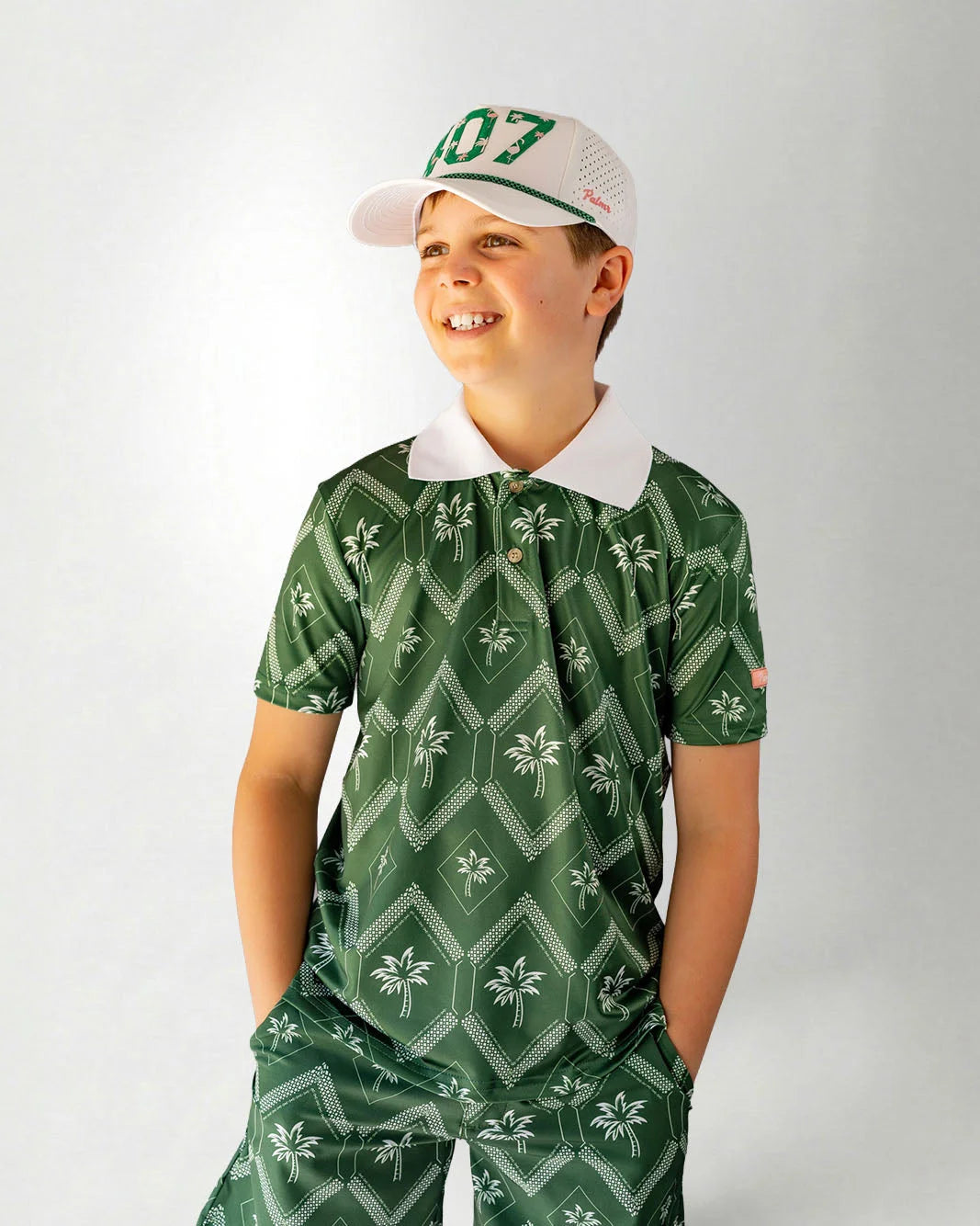 Child wearing a green patterned outfit with a white cap on a plain background