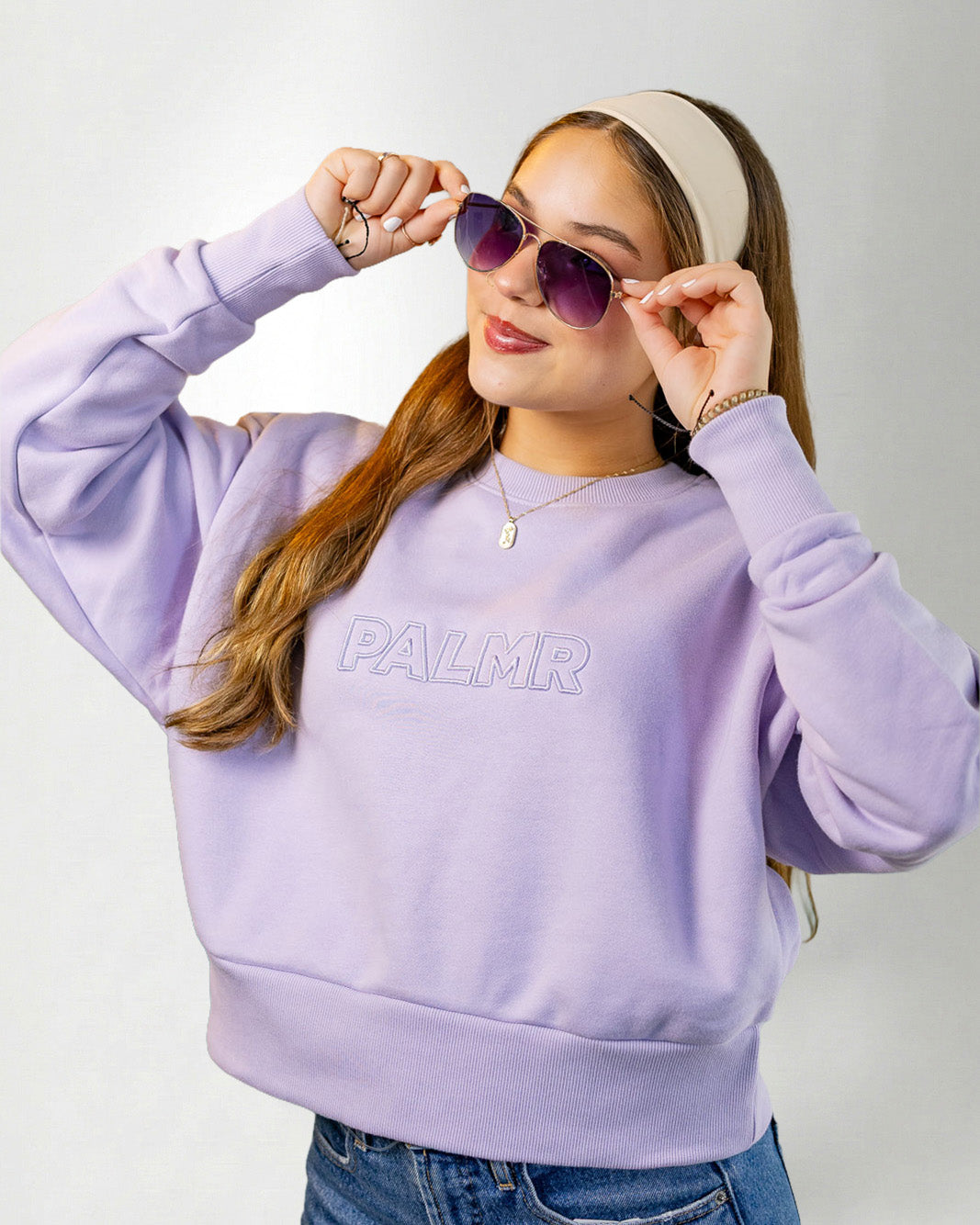 Woman wearing a lavender sweatshirt with 'PALM' branding, sunglasses, and a headband on a white background.
