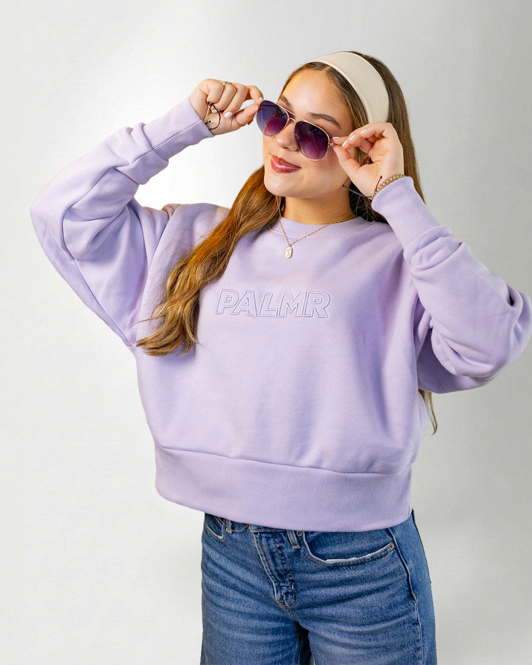 Woman wearing a lavender sweatshirt with 'PALM' branding, sunglasses, and a headband on a white background.