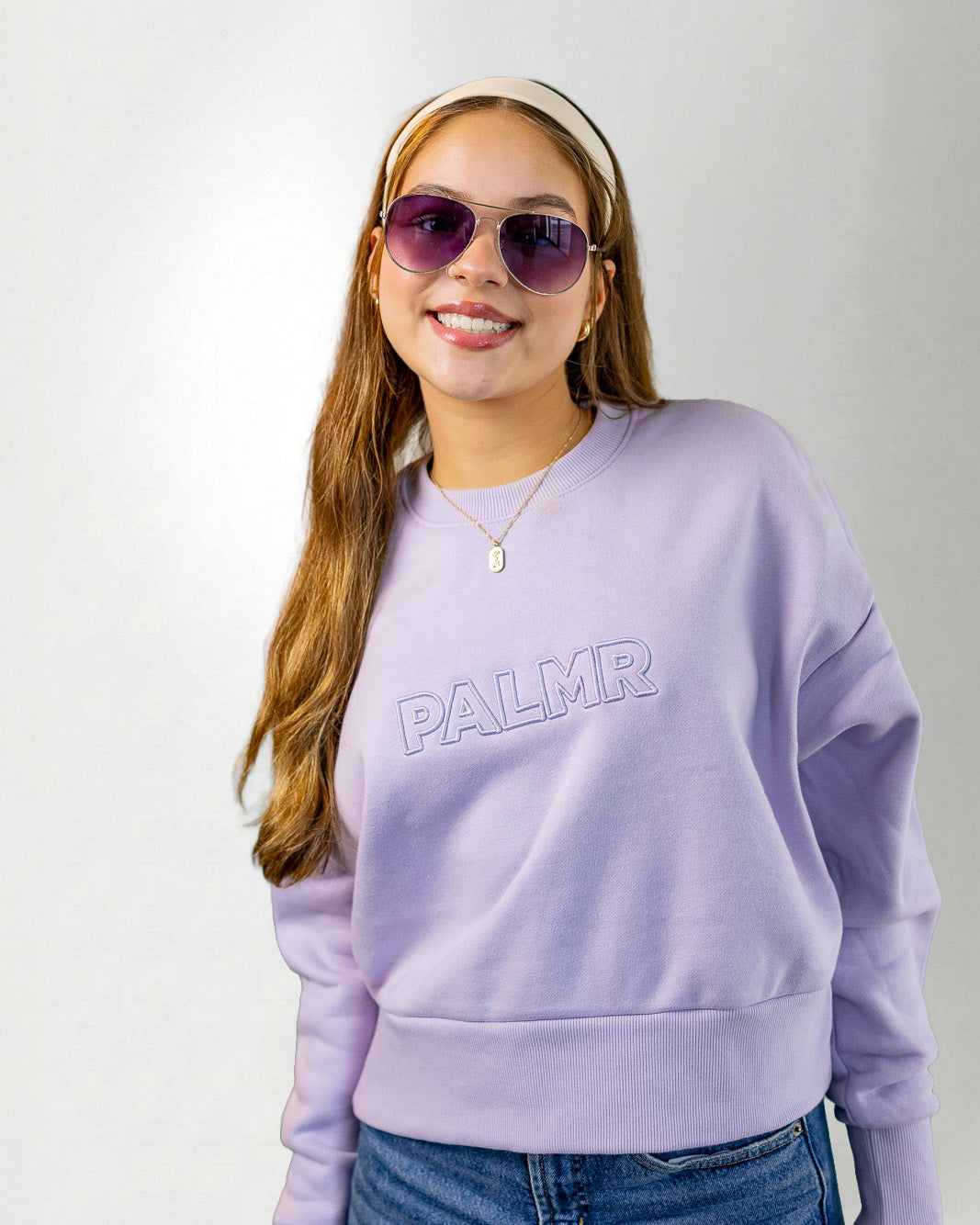 Woman wearing a lavender sweatshirt with 'PALM' branding, sunglasses, and a headband on a white background.
