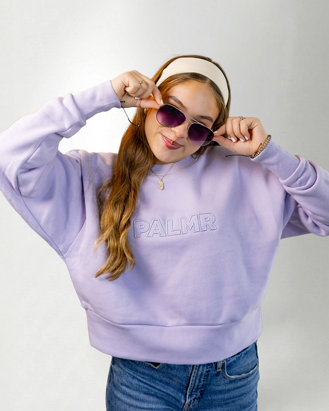 Woman wearing a lavender sweatshirt with 'PALM' branding, sunglasses, and a headband on a white background.