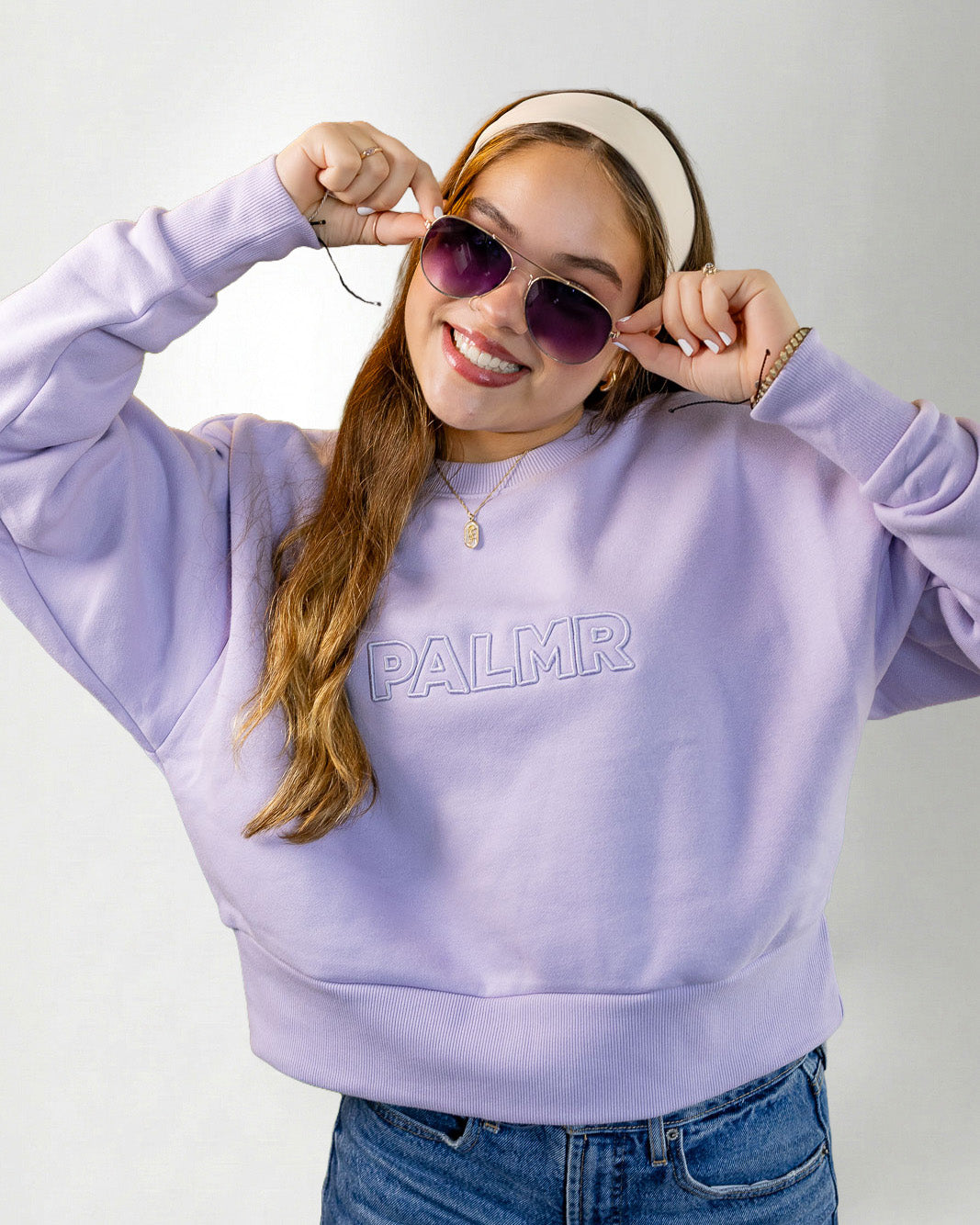 Woman wearing a lavender sweatshirt with 'PALM' branding, sunglasses, and a headband on a white background.
