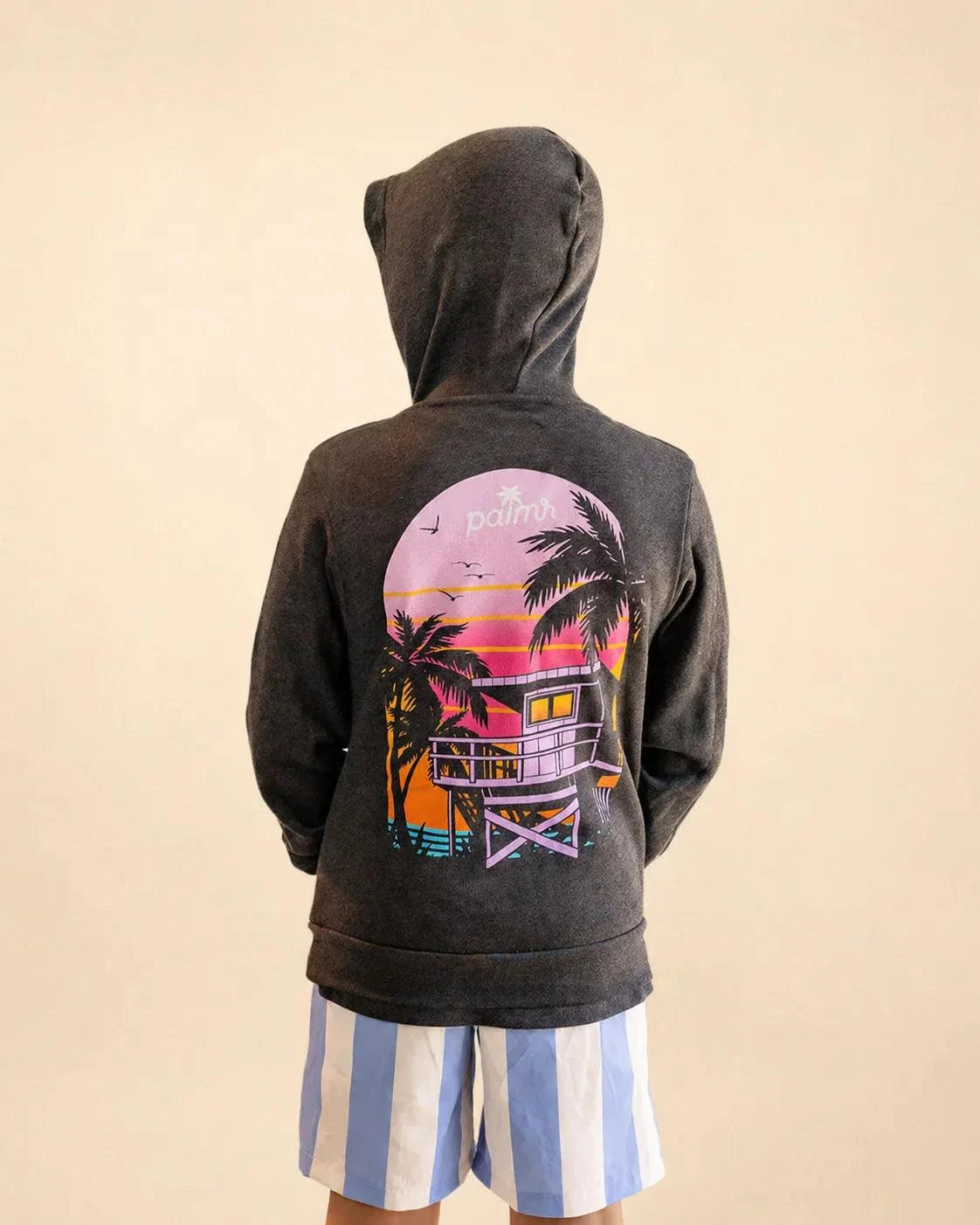 Child wearing The Evening Palm Kids Hoodie in dark heather grey with Palmr logo and sunset graphic