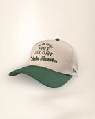 A baseball cap with a light beige crown and a dark green curved brim, featuring embroidered text on the front and side.