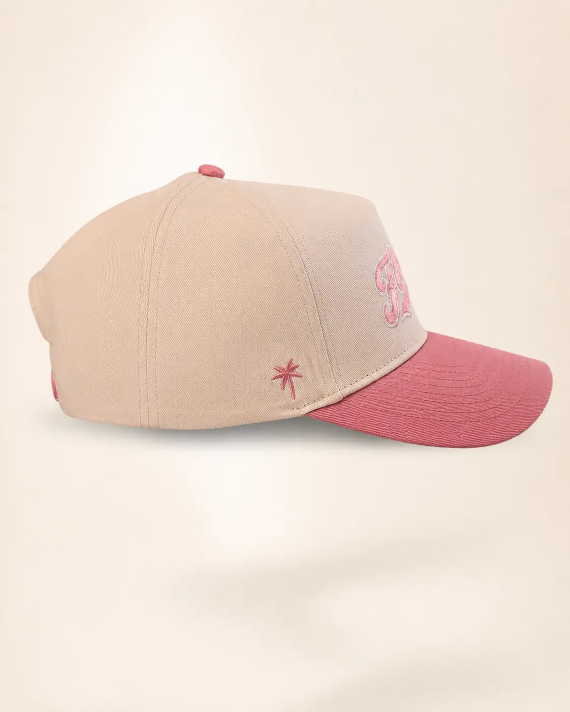 A structured baseball cap in beige with a dusty pink brim, featuring a pink embroidered star on the side and a pink embroidered monogram on the front.