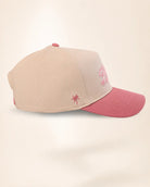 A structured baseball cap in beige with a dusty pink brim, featuring a pink embroidered star on the side and a pink embroidered monogram on the front.