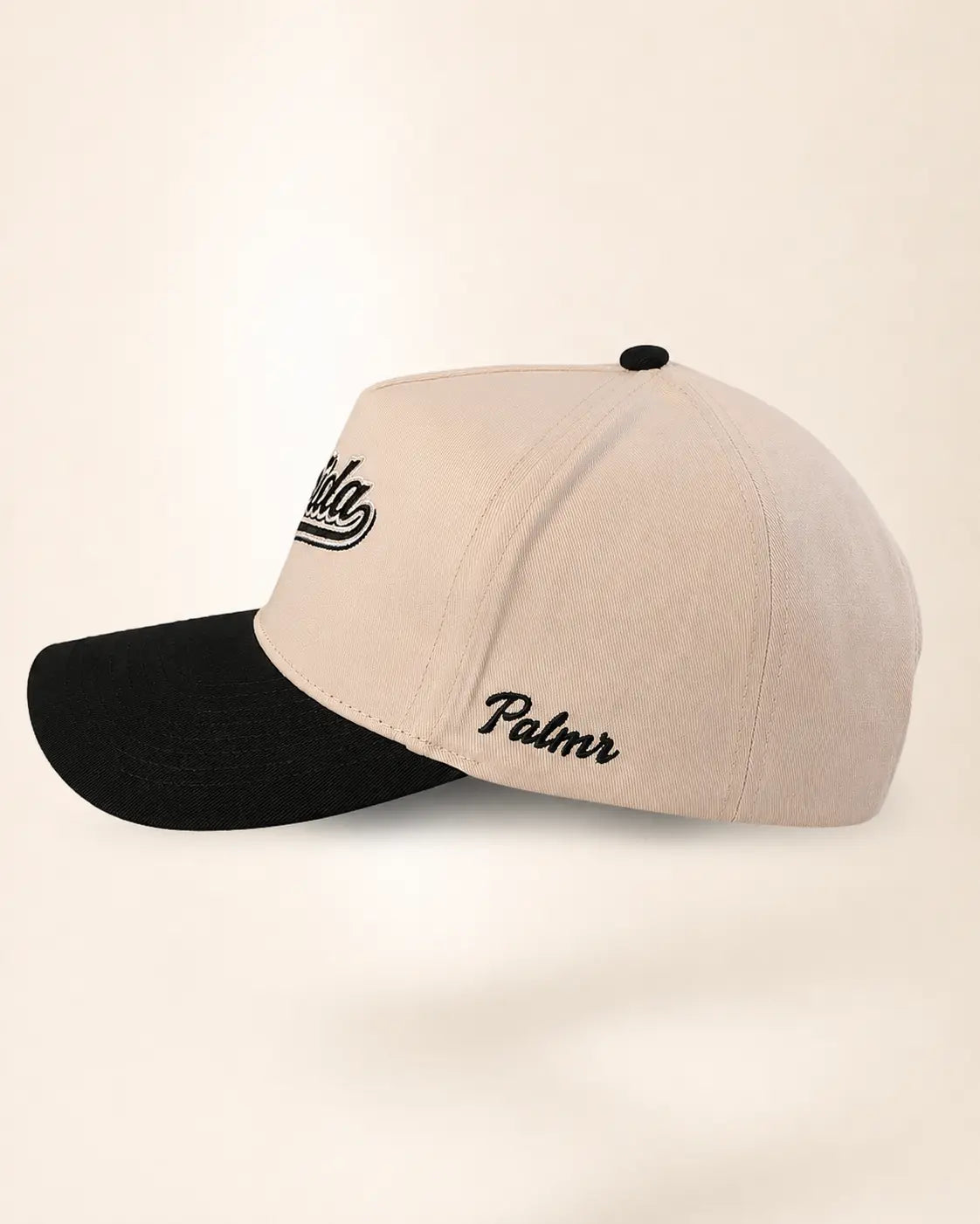 A structured baseball cap with a light beige crown and a black curved brim, featuring black embroidered script logos on the side, set against a plain cream background.