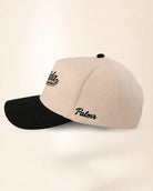 A structured baseball cap with a light beige crown and a black curved brim, featuring black embroidered script logos on the side, set against a plain cream background.
