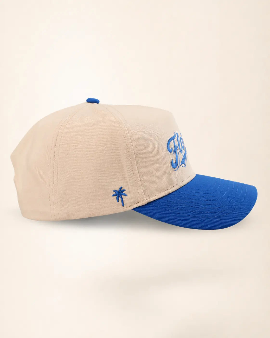A baseball cap with a cream-colored crown and a bright blue brim, featuring blue embroidered details including a palm tree and stylized lettering on the side.