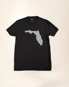 A black short-sleeve t-shirt with a gray silhouette graphic of the state of Florida on the front, laid flat against a light beige background.