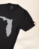 A close-up of a black short-sleeve t-shirt featuring a gray silhouette map of Florida with a small Palmr logo on the interior, shown against a light beige background.