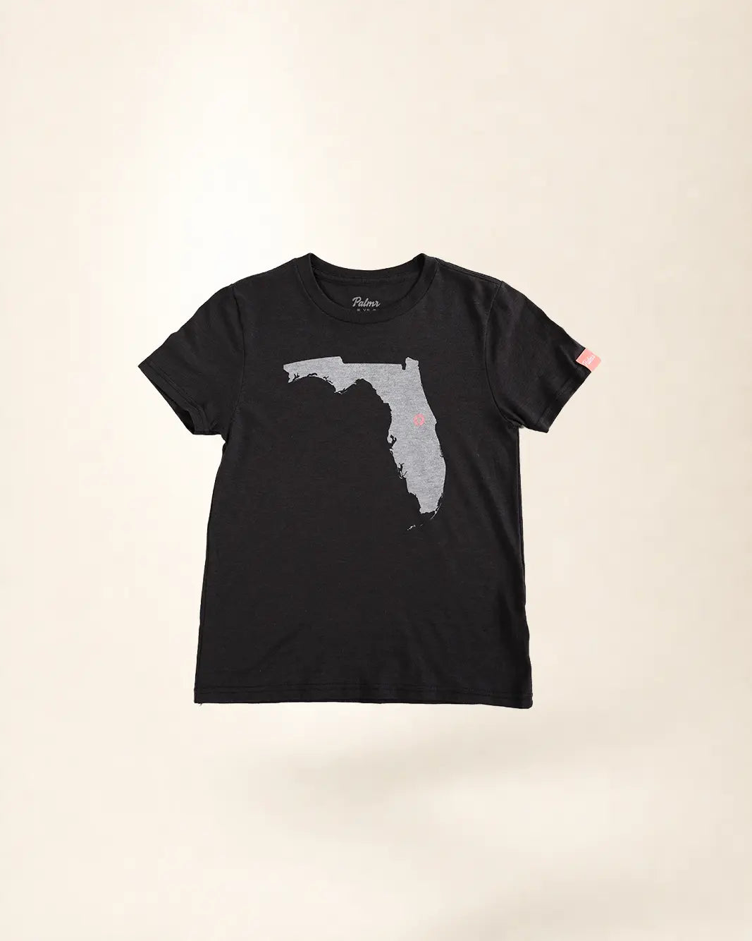 A black short-sleeve t-shirt with a gray outline graphic of the state of Florida on the front, featuring a small pink dot on the southeast coast, displayed against a light beige background.