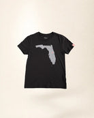 A black short-sleeve t-shirt with a gray outline graphic of the state of Florida on the front, featuring a small pink dot on the southeast coast, displayed against a light beige background.