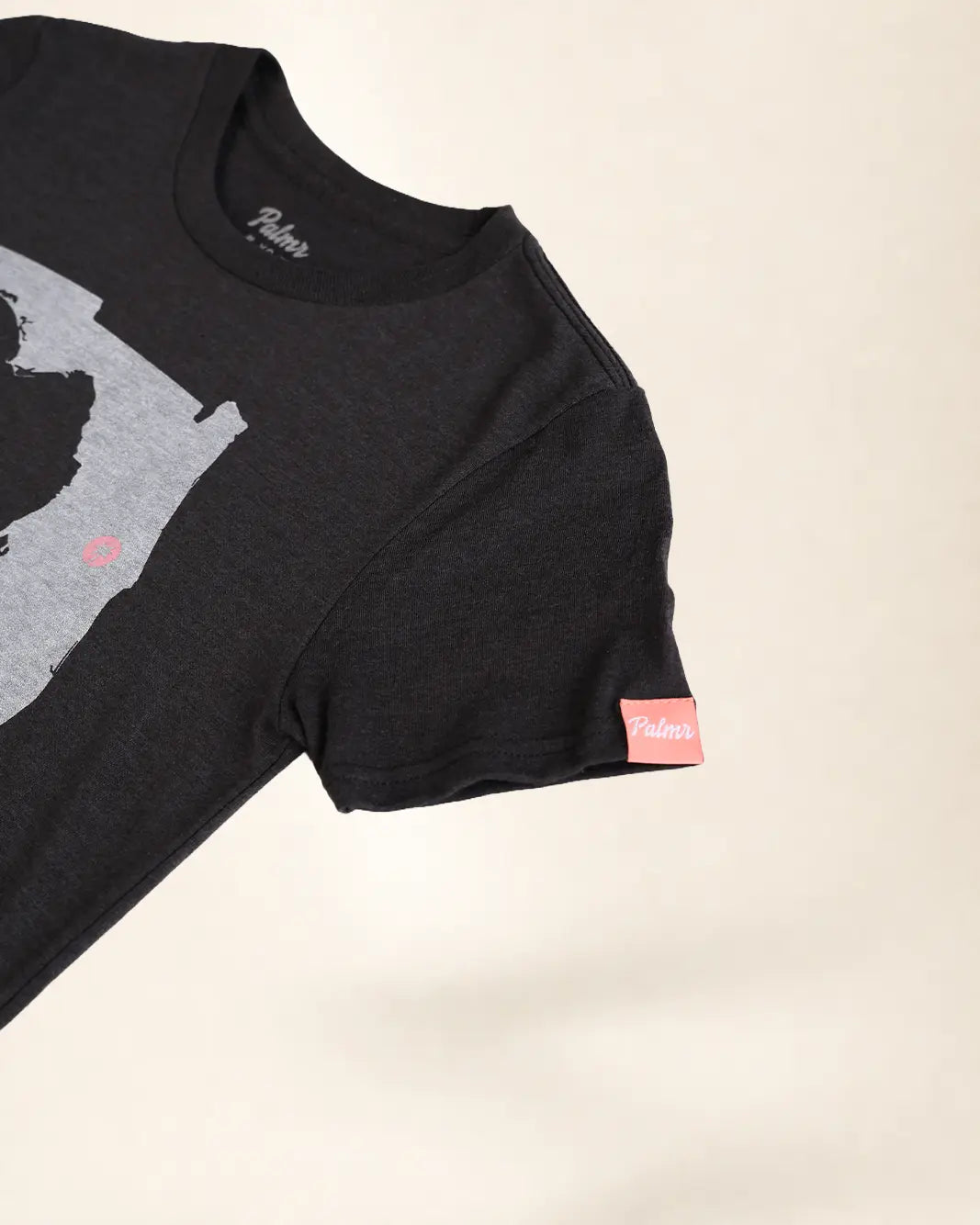 A close-up of a black short-sleeve t-shirt with a gray graphic print on the front and a small pink brand tag on the sleeve, set against a plain light beige background.