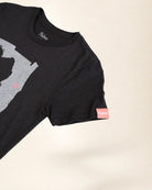 A close-up of a black short-sleeve t-shirt with a gray graphic print on the front and a small pink brand tag on the sleeve, set against a plain light beige background.