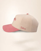 A structured baseball cap in light beige with a contrasting dusty pink curved brim, featuring pink embroidered script text on the front and side.