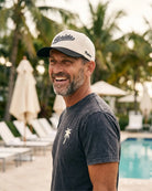 A smiling man wearing a white and navy Florida cap with a dark gray t-shirt stands by a swimming pool with palm trees in the background.