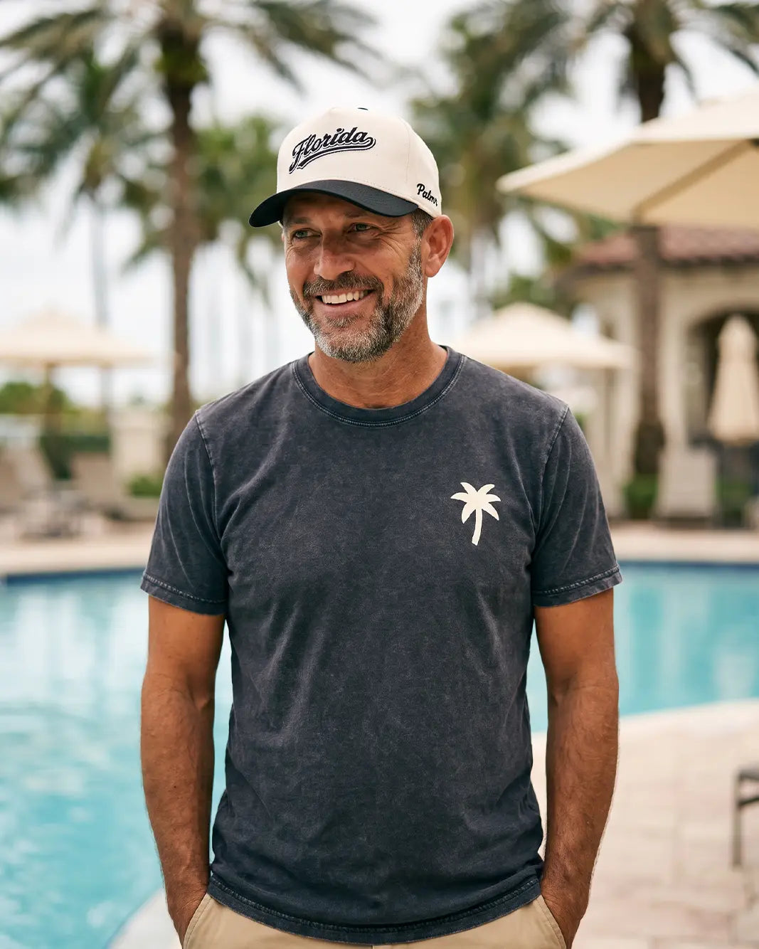 A smiling man standing by a swimming pool wearing a dark charcoal gray t-shirt with a small white palm tree logo on the left chest, paired with a white and navy Florida baseball cap.