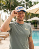 A smiling man standing by a pool wearing a gray t-shirt with 'Palmer' embroidered in gold on the chest, and a white and blue 'Florida' baseball cap.