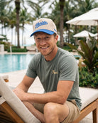 A smiling man sitting on a poolside lounge chair wearing a beige and blue Florida trucker hat, a muted green t-shirt with gold embroidery, and khaki shorts, with palm trees and resort umbrellas in the background.