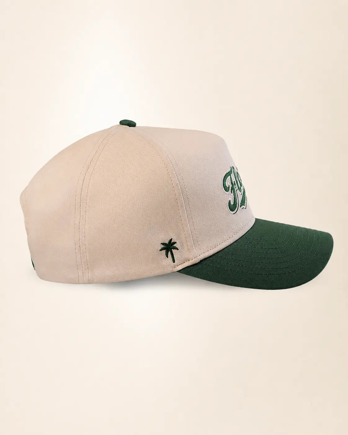 A baseball cap with a light beige crown and a dark green curved brim, featuring green embroidered script on the side and a small green palm tree logo.