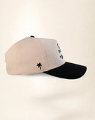 A baseball cap with a light beige crown and a black curved brim, featuring a small black palm tree logo on the side and black embroidered text on the front.