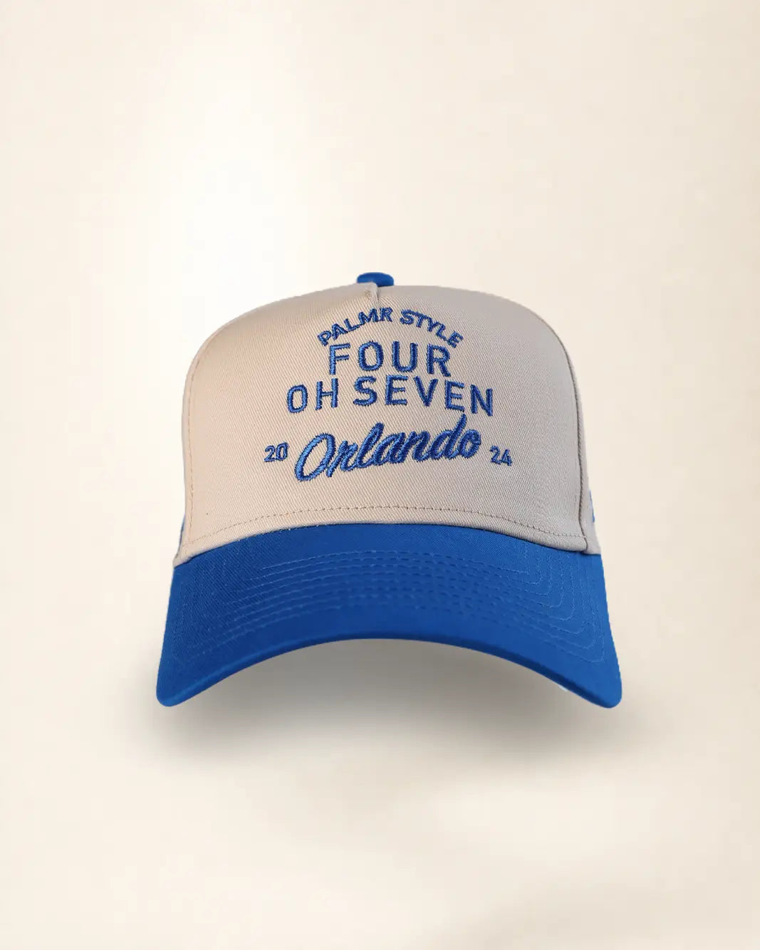 A two-tone baseball cap with a cream-colored front panel and brim, and a royal blue curved brim and top button. The front panel features blue embroidered text reading 'PALMR STYLE', 'FOUR OH SEVEN', and 'Orlando 26'.