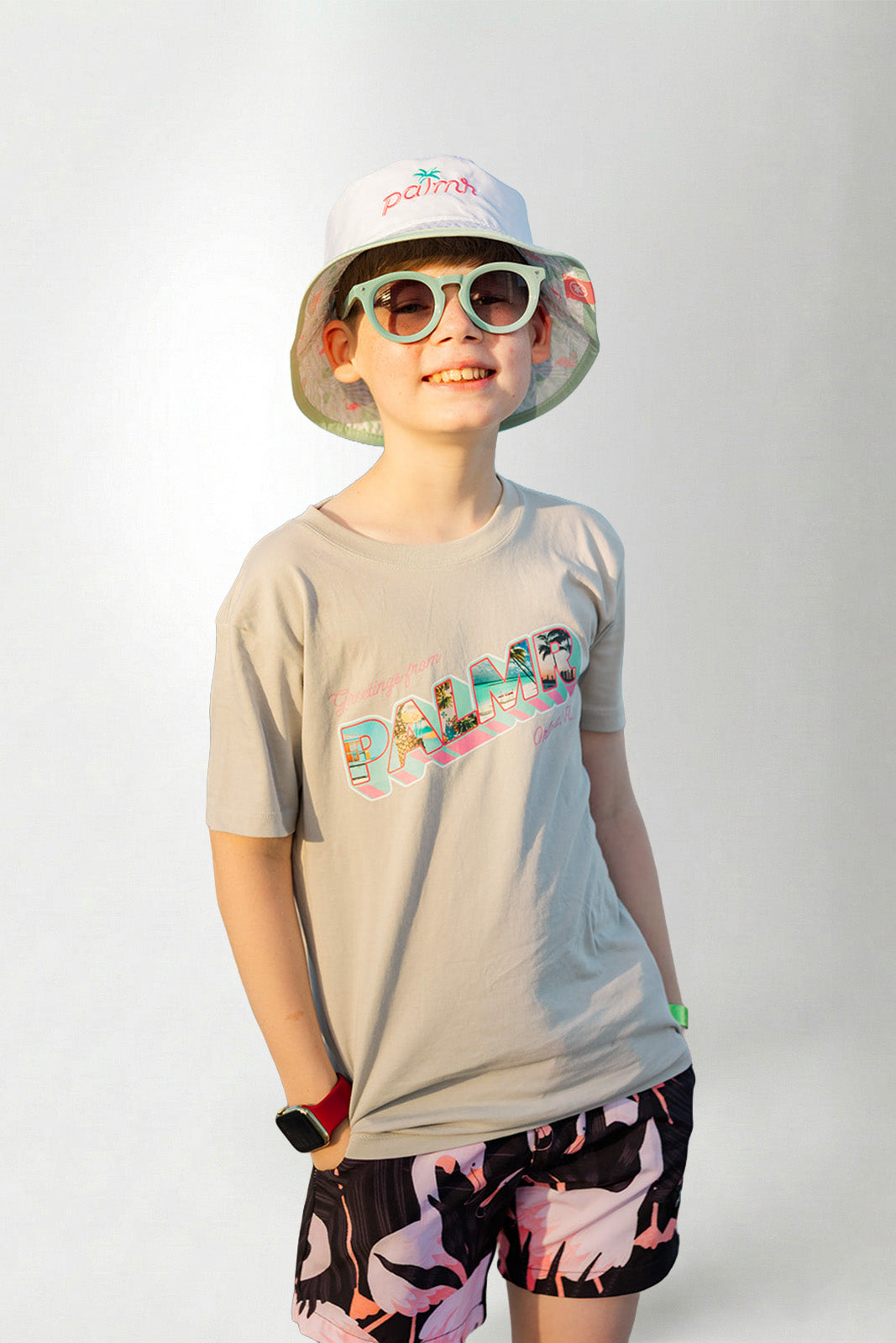 Person wearing a light gray t-shirt with colorful text, black shorts with pink designs, and a white bucket hat with green rim on a plain background.