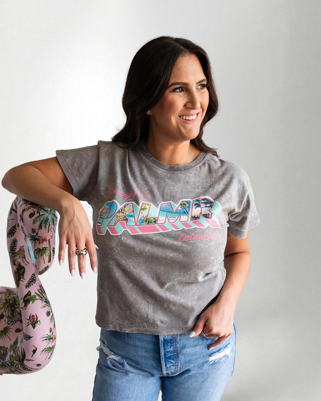 Woman wearing a gray t-shirt with colorful text and blue jeans, posing against a plain background.