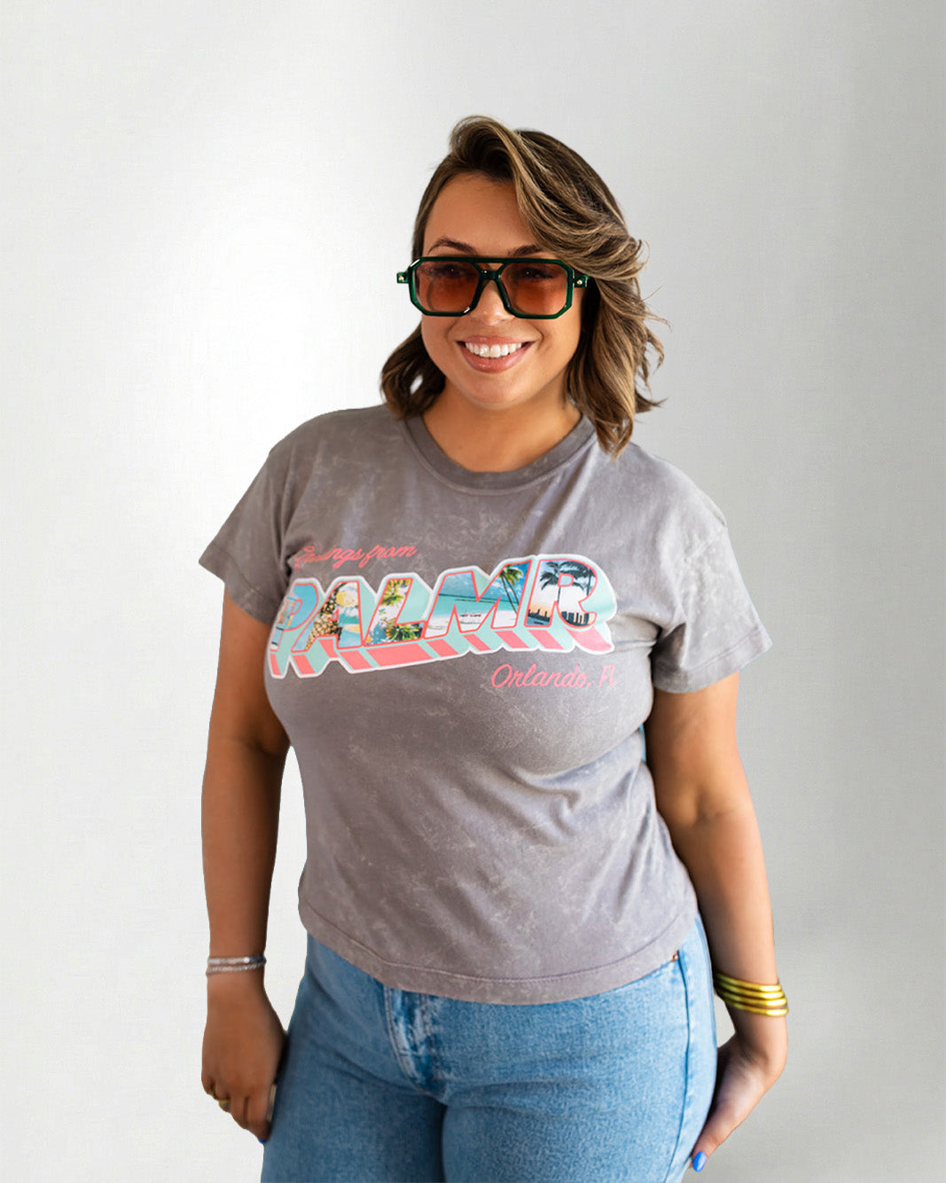 Woman wearing a gray t-shirt with colorful text and blue jeans, posing against a plain background.