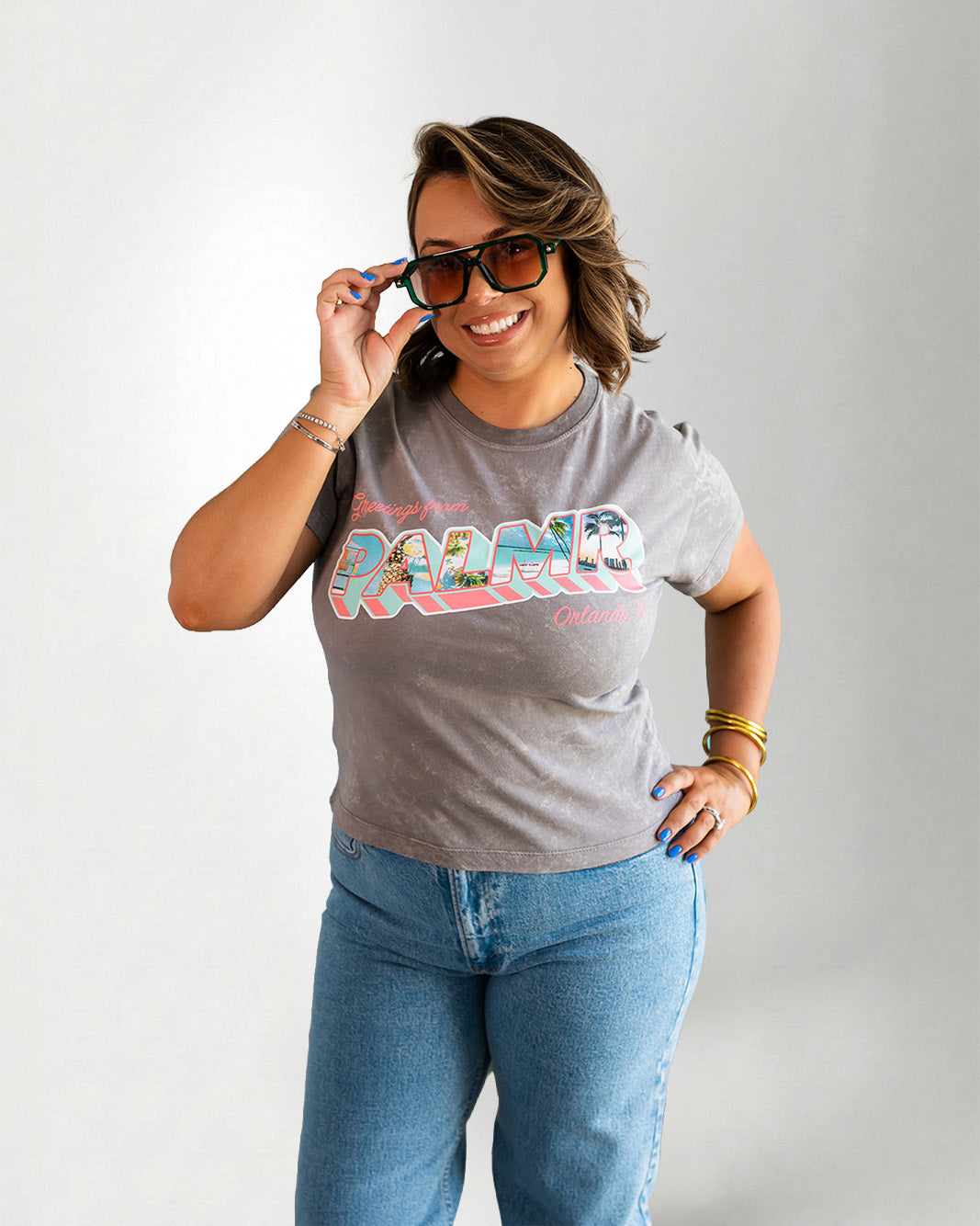Woman wearing a gray t-shirt with colorful text and blue jeans, posing against a plain background.
