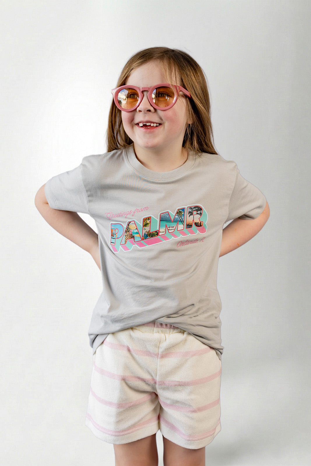 Young girl wearing a gray t-shirt with 'PALMIR' print and striped shorts on a white background
