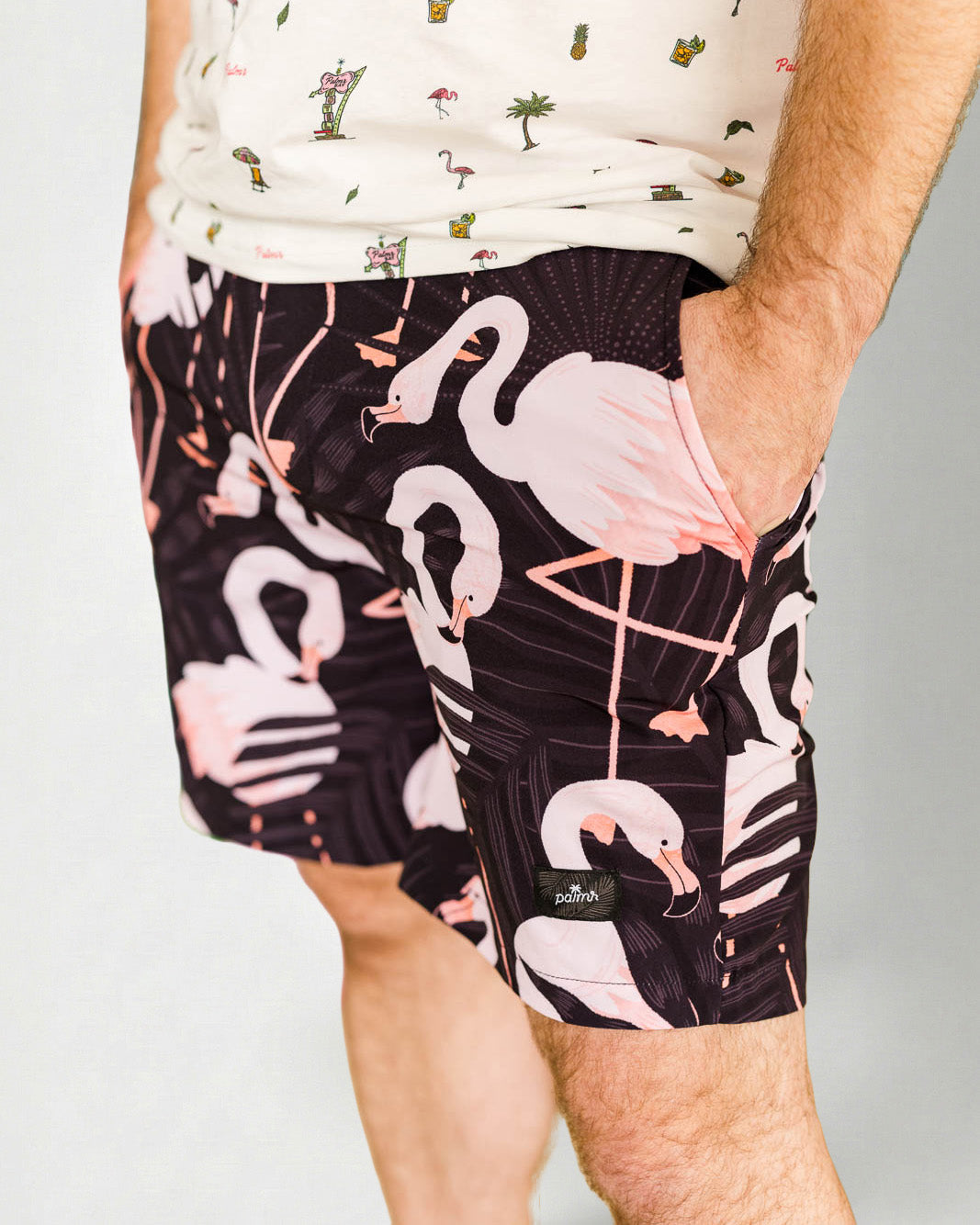 Person wearing a white shirt with a palm tree pattern and black shorts with pink flamingo pattern on a plain background