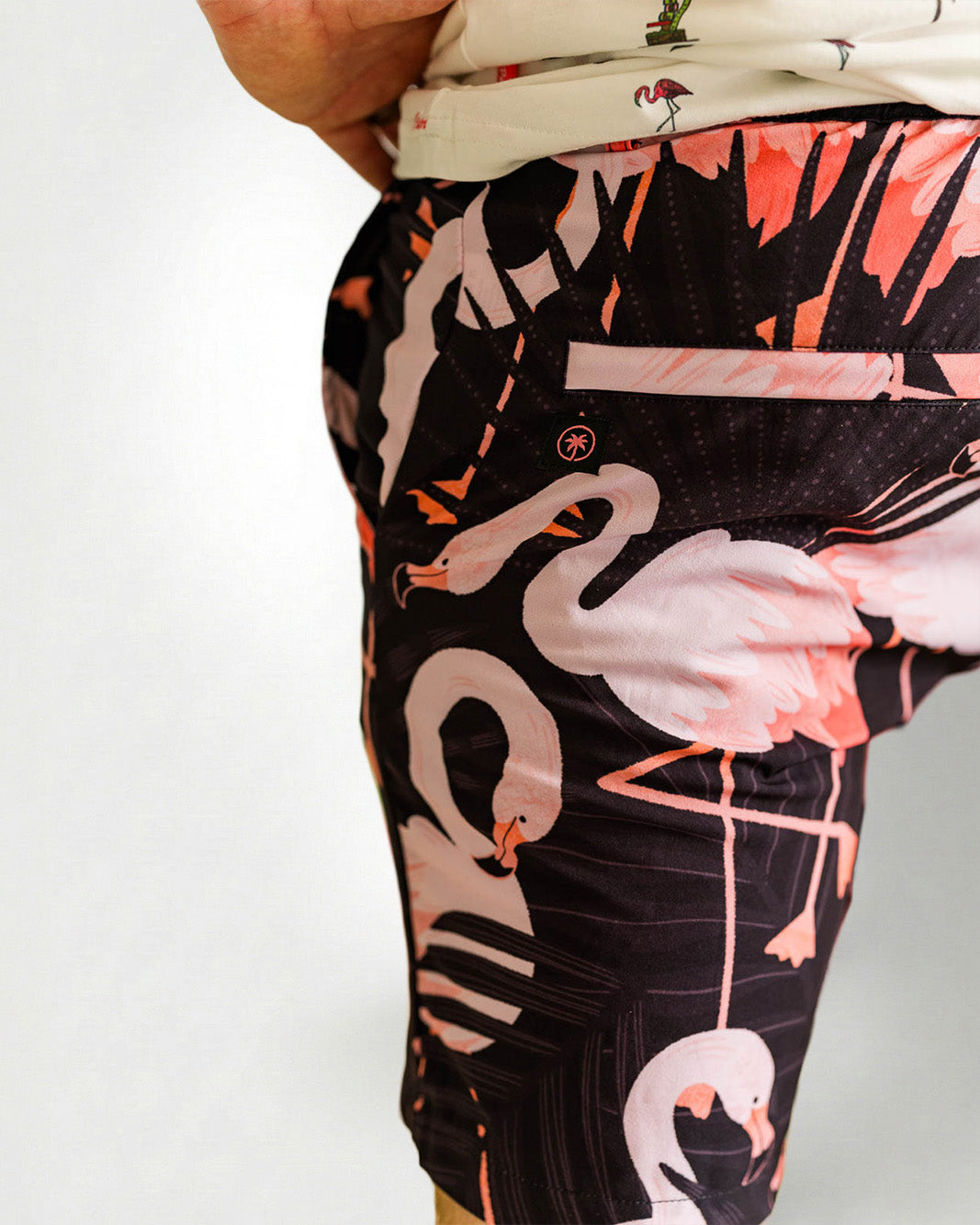 Person wearing a white shirt with a palm tree pattern and black shorts with pink flamingo pattern on a plain background