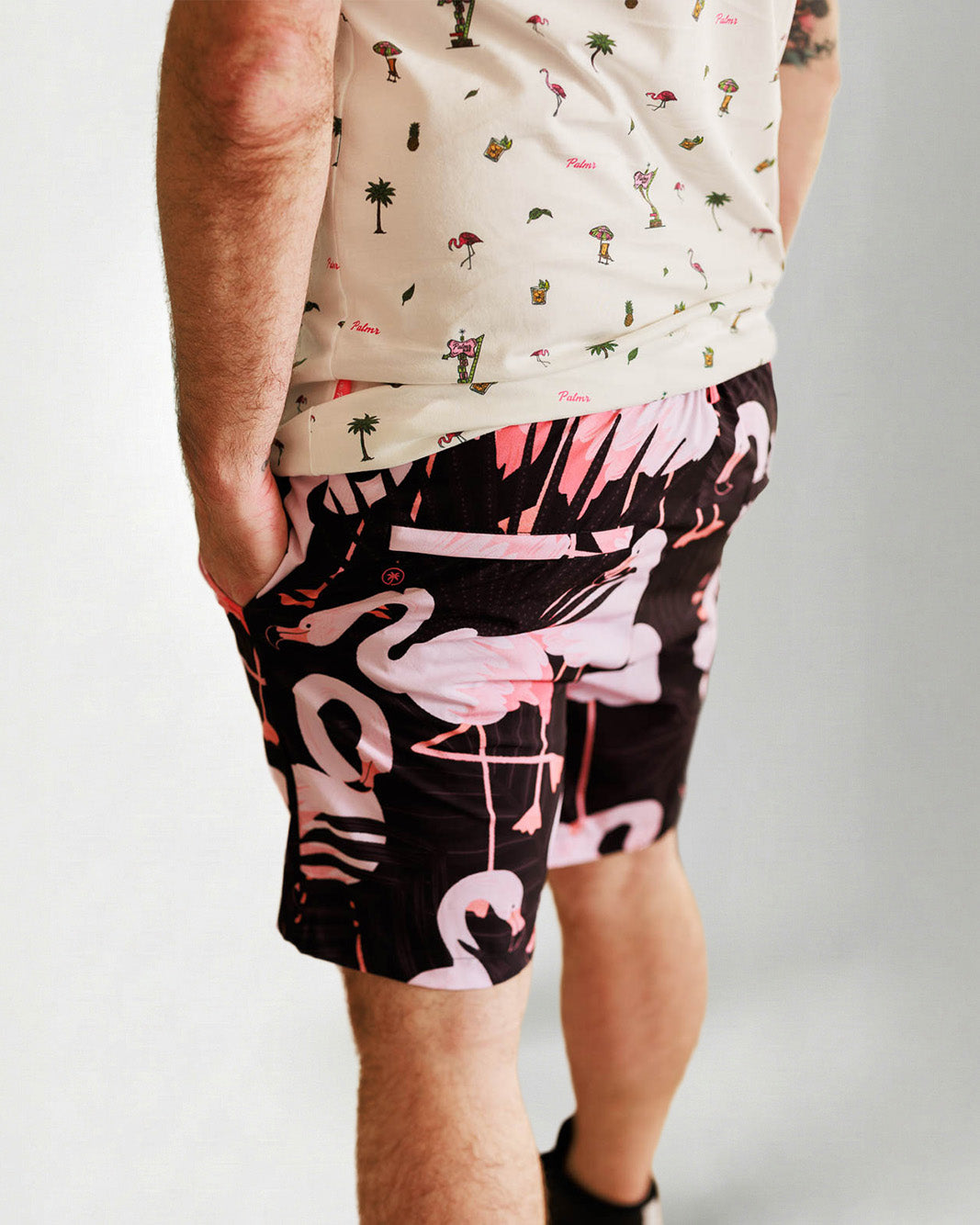 Person wearing a white shirt with a palm tree pattern and black shorts with pink flamingo pattern on a plain background