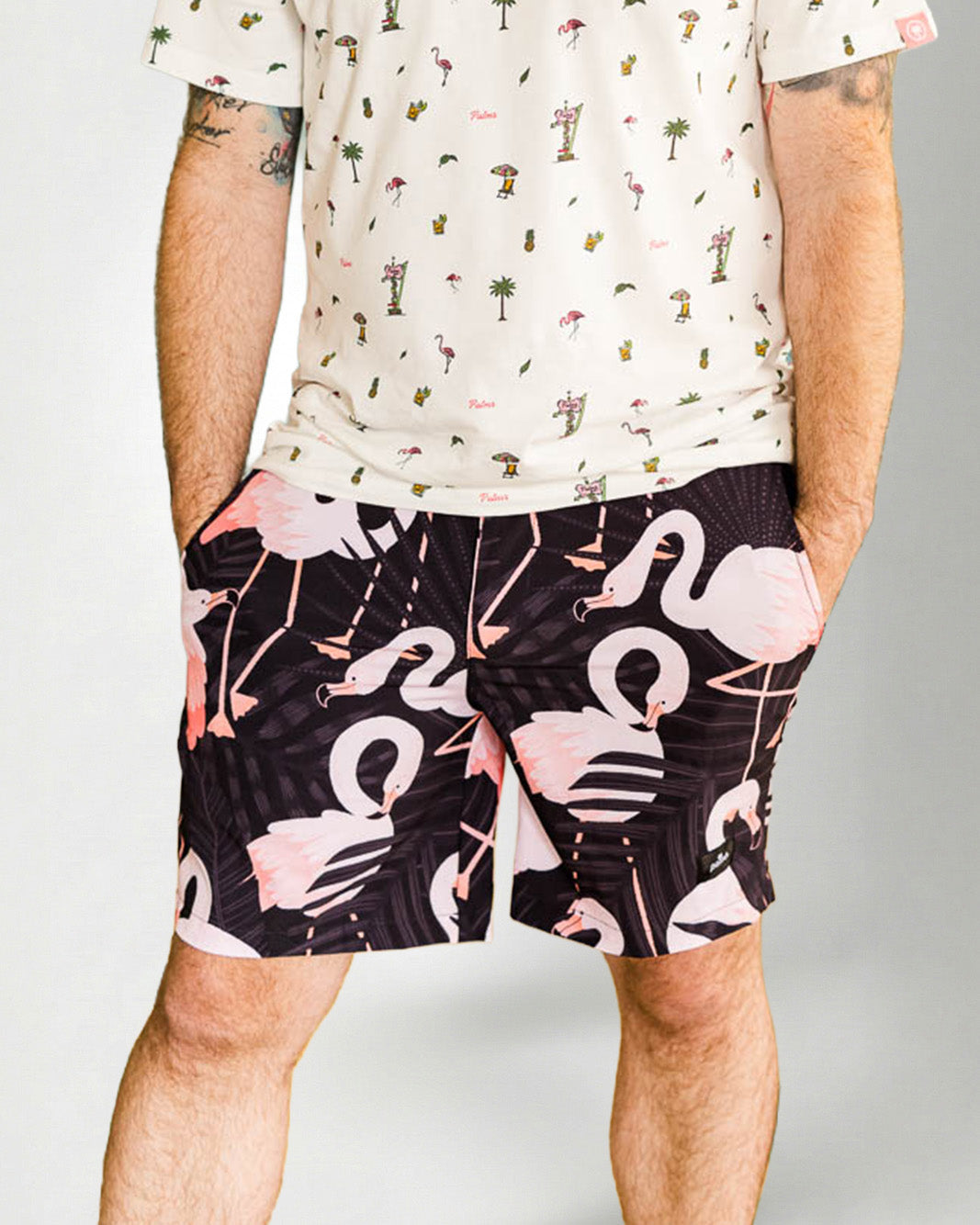 Person wearing a white shirt with a palm tree pattern and black shorts with pink flamingo pattern on a plain background