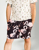 Person wearing a white shirt with a palm tree pattern and black shorts with pink flamingo pattern on a plain background