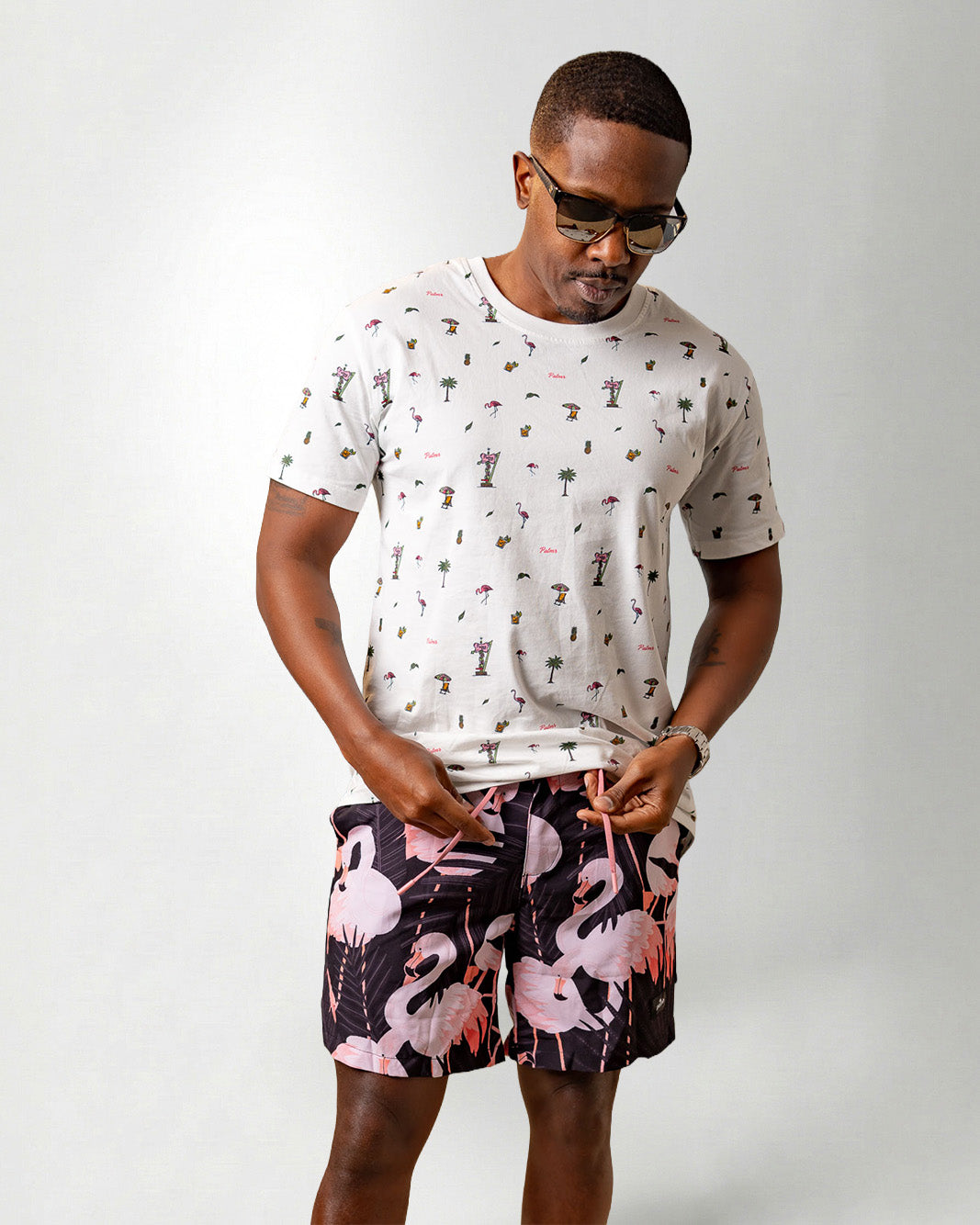 Person wearing a white shirt with a palm tree pattern and black shorts with pink flamingo pattern on a plain background